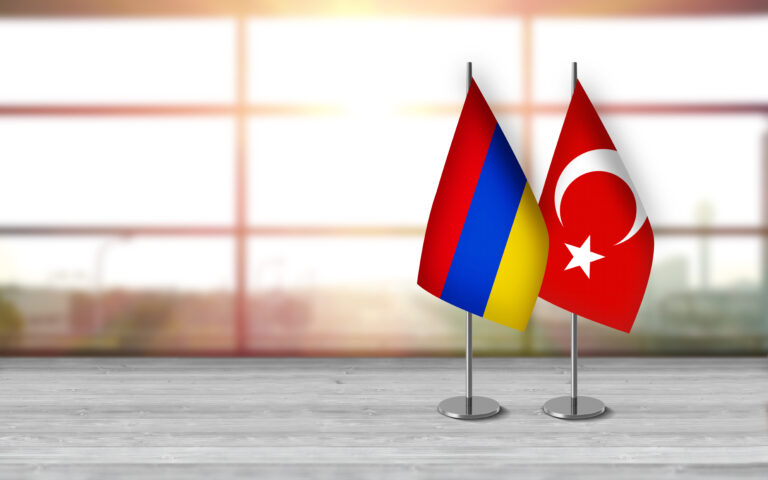 Armenian and turkish 3D desk flags are standing together on a desk in front of sunny background with large copy space.
