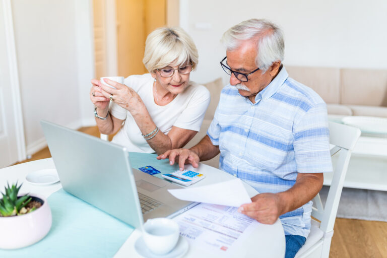 Happy middle aged husband and wife sitting at table with laptop and paper bills, calculating domestic incomes together at home.
