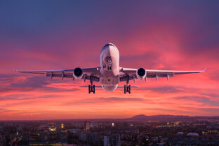 Airplane is flying in colorful sky over city at night. Landscape with passenger airplane, skyline, purple sky with red and pink clouds at dusk. Aircraft is landing at sunset. Aerial view of plane