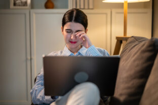 Exhausted Asian young woman hurt eye while using laptop in dark room. Attractive girl sitting on sofa feeling visual fatigue and eye strain tired from overwork and massage dry irritable eye at home.