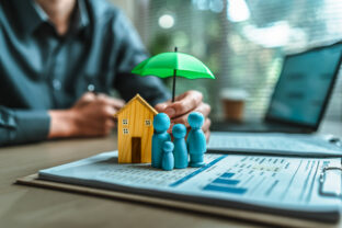 A wooden doll and wood house under an umbrella holding by agent represent insurance . house Family , protection, life.