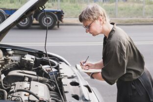 Middle aged woman holding a notepad stands in front of a car with the hood up