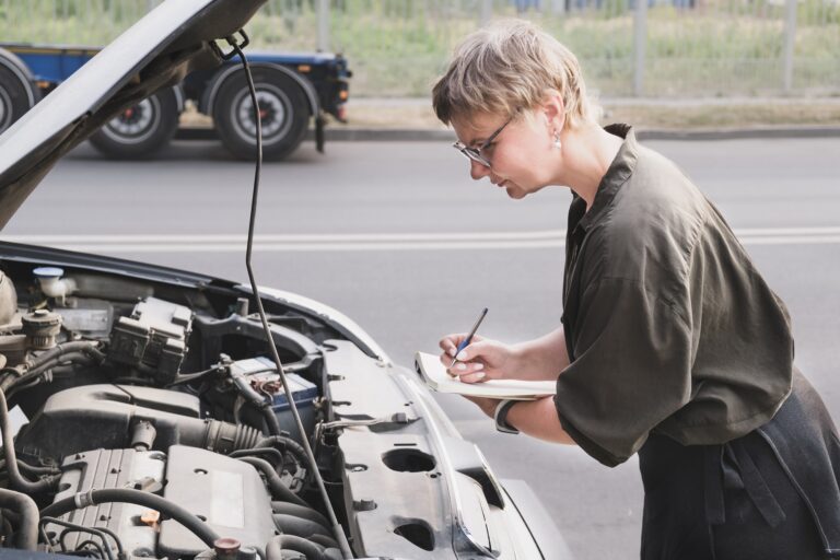 Middle aged woman holding a notepad stands in front of a car with the hood up
