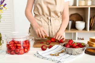 Woman cutting fresh berries for preparing sweet strawberry jam at table in kitchen