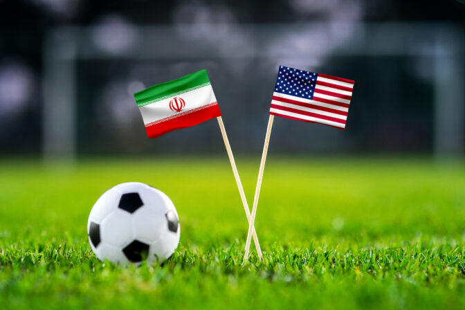 Iran vs. USA, Al Thumama, Football match wallpaper, Handmade national flags and soccer ball on green grass. Football stadium in background. Black edit space.