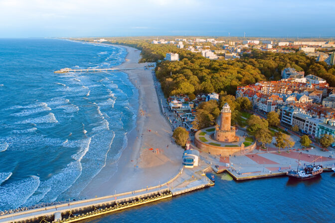 Drone photo captures Koobrzeg's maritime charm, featuring the iconic lighthouse, cerulean sea, turbulent waves, a distant pier, and autumnal hues on the trees.