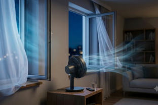Stolný ventilátor umiestnený na stole meter od okna, fúkajúci teplý vzduch von do letnej noci, zatiaľ čo závesy sa jemne hýbu v prievane.