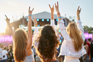 Group of female friends enjoying live music on sunny beach festival. Girls dance, celebrate with hands up in summer vibe. Casual fun, youth culture, outdoor event by sea. Friendship, lifestyle moment.