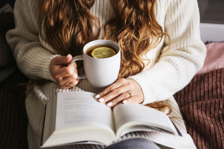 Cozy scene of woman sitting with cup of hot tea and reading book on bed.