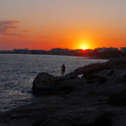 A tourist stands on the rocks at the 'Love' natural bridge during the sunset in the resort of Ayia Napa in the eastern Mediterranean island of Cyprus, Sunday, April 23, 2023. Ayia Napa is destination for thousands of British and European tourists every summer