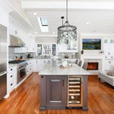 Long kitchen island with storage and dining space along with white cabinetry and big fridge aside the living room with cozy sofa and fireplace.jpg